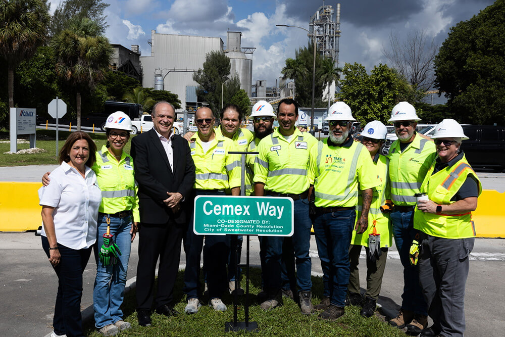 A group of people, including workers in safety gear and two people in business attire, stand outdoors by a green Cemex Way street sign, smiling for a photo with an industrial facility in the background.