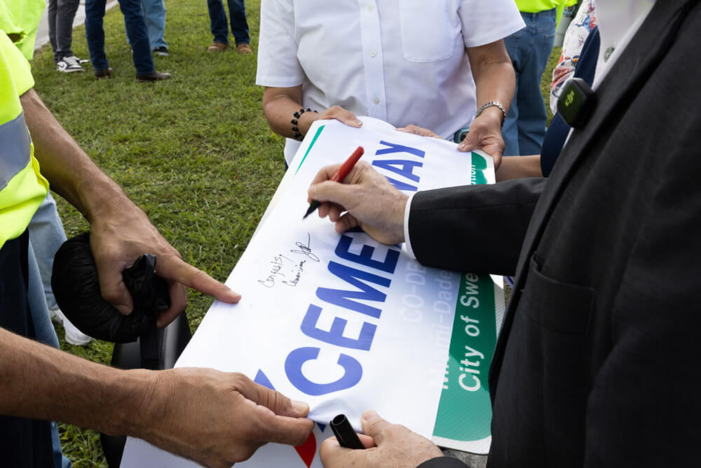 Several people hold a sign as a person in a suit signs it with a black pen. The group is outdoors on grass, and only their hands and torsos are visible.