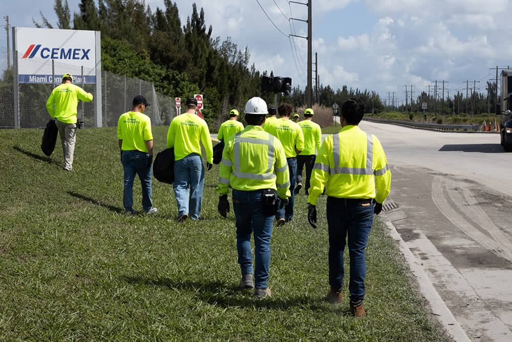 A group of workers in bright yellow safety shirts and hard hats walk along a grassy roadside near a CEMEX sign, with power lines and cloudy skies in the background.