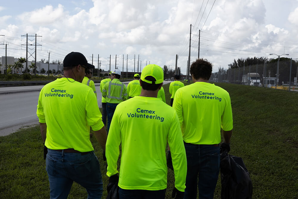 A group of people in bright yellow Cemex Volunteering shirts walk along a grassy roadside, collecting trash, with power lines and cloudy skies in the background.