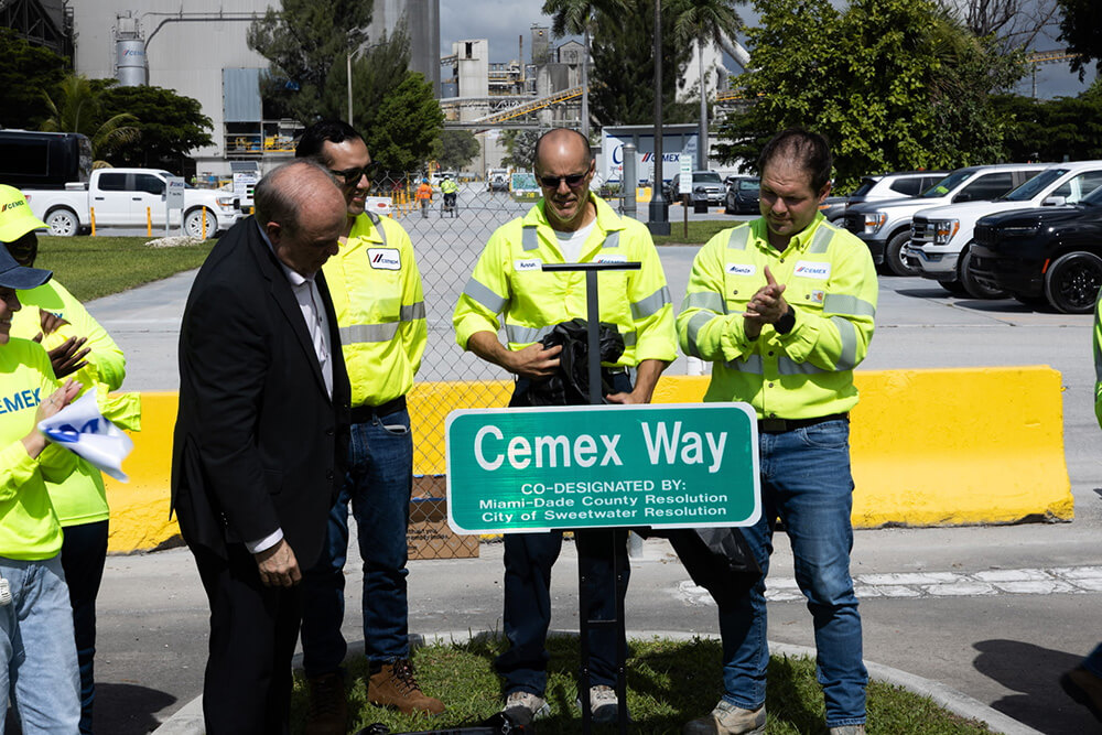 Four people wearing safety vests stand around a new green road sign that reads Cemex Way, during a daytime unveiling ceremony outdoors near a construction site.
